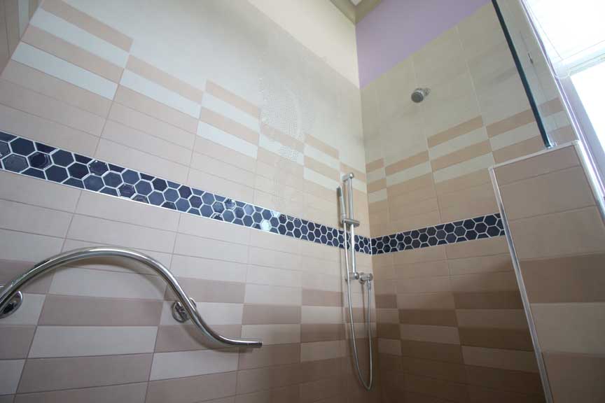 Close up of Handicapped accessible shower with curved grab bar, grab bar hand shower and no shower valves. The wall is tiled in a gradation of the sunrise.